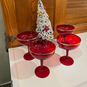 May include: Four red stemmed champagne glasses with gold rims. The glasses are arranged in a semi-circle on a white surface. A small, snowy artificial Christmas tree is in the background.
