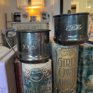 Peut inclure: Deux tasses en argent avec des initiales gravées, F.J.S. et E.R.H., sont empilées sur des livres anciens. Les livres ont des inscriptions dorées sur leurs dos, dont "The Girls Own Annual" et "The Literature of America".