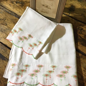 May include: Two white pillowcases with scalloped edges and embroidered green and orange flowers. The pillowcases are on a wooden surface with a vintage photo of a baby in a frame.