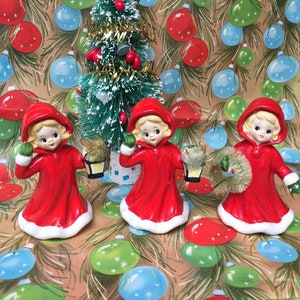 Op de afbeelding: Drie vintage porseleinen beeldjes van meisjes die rode gekapte jurken dragen en kerstversiering vasthouden. De beeldjes staan voor een kleine kunstkerstboom met gouden tinsel en rode kerstballen. De achtergrond is een bruin en groen inpakpapier met een herhalend patroon van kerstballen.