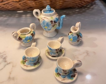 Puede incluir: Un juego de té en miniatura con diseños florales azules y marrones. El juego incluye una tetera, una jarra de crema, un azucarero y cuatro tazas de té y platillos.