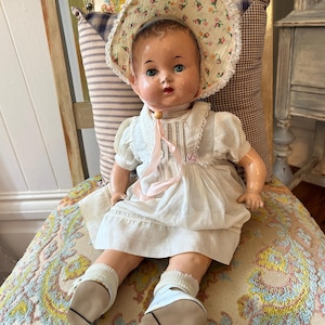 May include: A vintage doll with a painted face, blue eyes, and rosy cheeks. The doll wears a white dress with short sleeves, a bonnet featuring a small floral pattern with lace trim, white socks, and white shoes tied with a light pink ribbon.