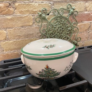 May include: A white ceramic casserole dish with a green rim and a Christmas tree design. The dish is sitting on a black hob.