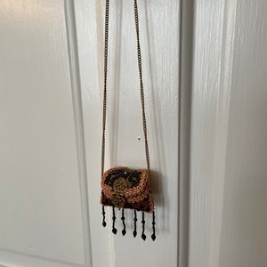 May include: A small, ornate, brown and black purse with a gold clasp and black beaded fringe hanging from a gold chain.