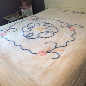 May include: A white crocheted bedspread with a blue and pink floral design. The bedspread has a raised texture and is made of a soft, fluffy material.