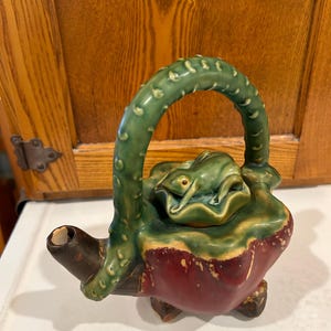May include: A green ceramic teapot with a frog on the lid and a vine-like handle. The teapot is decorated with a red glaze and has a rustic look.