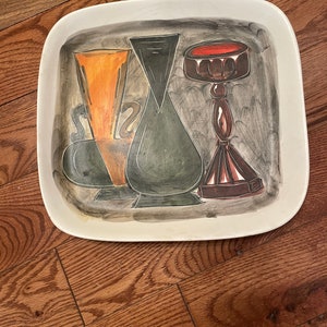 May include: A white ceramic serving tray with a hand-painted design of three stylized vases. The vases are painted in shades of black, green, orange, and brown. The tray has a square shape with rounded corners.