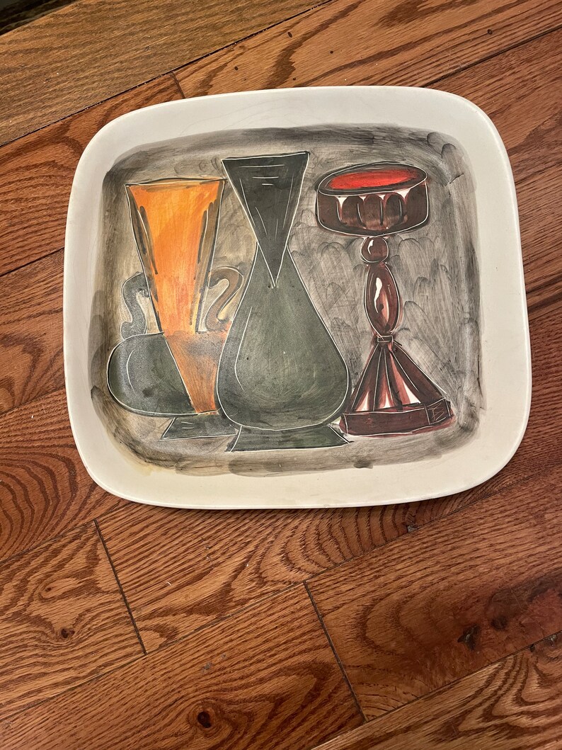 May include: A white ceramic serving tray with a hand-painted design of three stylized vases. The vases are painted in shades of black, green, orange, and brown. The tray has a square shape with rounded corners.