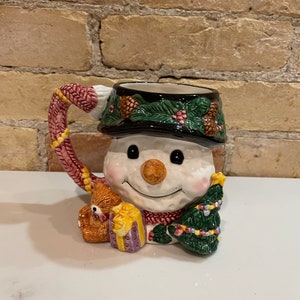 Könnte beinhalten: Eine Keramik-Schneemann-Tasse mit einem rot-weißen Schal, die einen kleinen Teddybären und einen Weihnachtsbaum hält. Die Tasse hat einen schwarzen Rand und einen grün-braunen Hut mit Tannenzapfen.