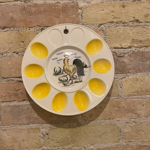 Puede incluir: Un plato de huevos de cerámica blanca con un diseño de soporte para huevos amarillo. El plato presenta una ilustración de gallo en el centro.
