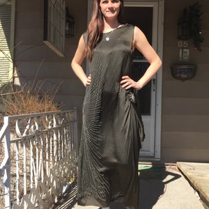 May include: A woman wearing a long, dark green, sequined dress with a silver necklace and a silver brooch. She is standing on a porch with a white railing and a wooden door behind her.