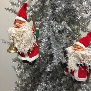 Puede incluir: Dos adornos de Papá Noel rojos y blancos colgados de un árbol de Navidad de oropel plateado. Los Papás Noel sostienen un palo y una campana.