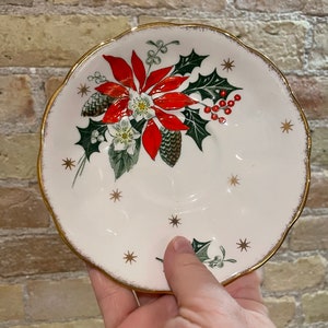 May include: White ceramic saucer with a gold rim and a Christmas-themed design featuring a red poinsettia, green holly leaves, and gold stars.