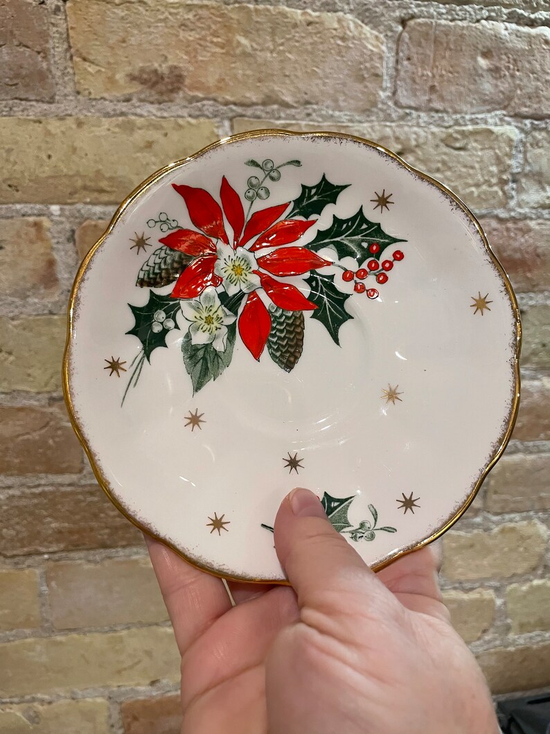 May include: White ceramic saucer with a gold rim and a Christmas-themed design featuring a red poinsettia, green holly leaves, and gold stars.