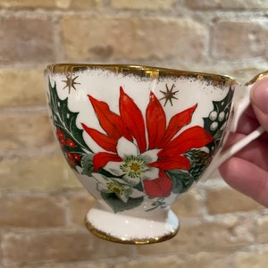 May include: White ceramic teacup with a gold rim and a Christmas-themed design featuring red poinsettias, green holly, and white flowers.