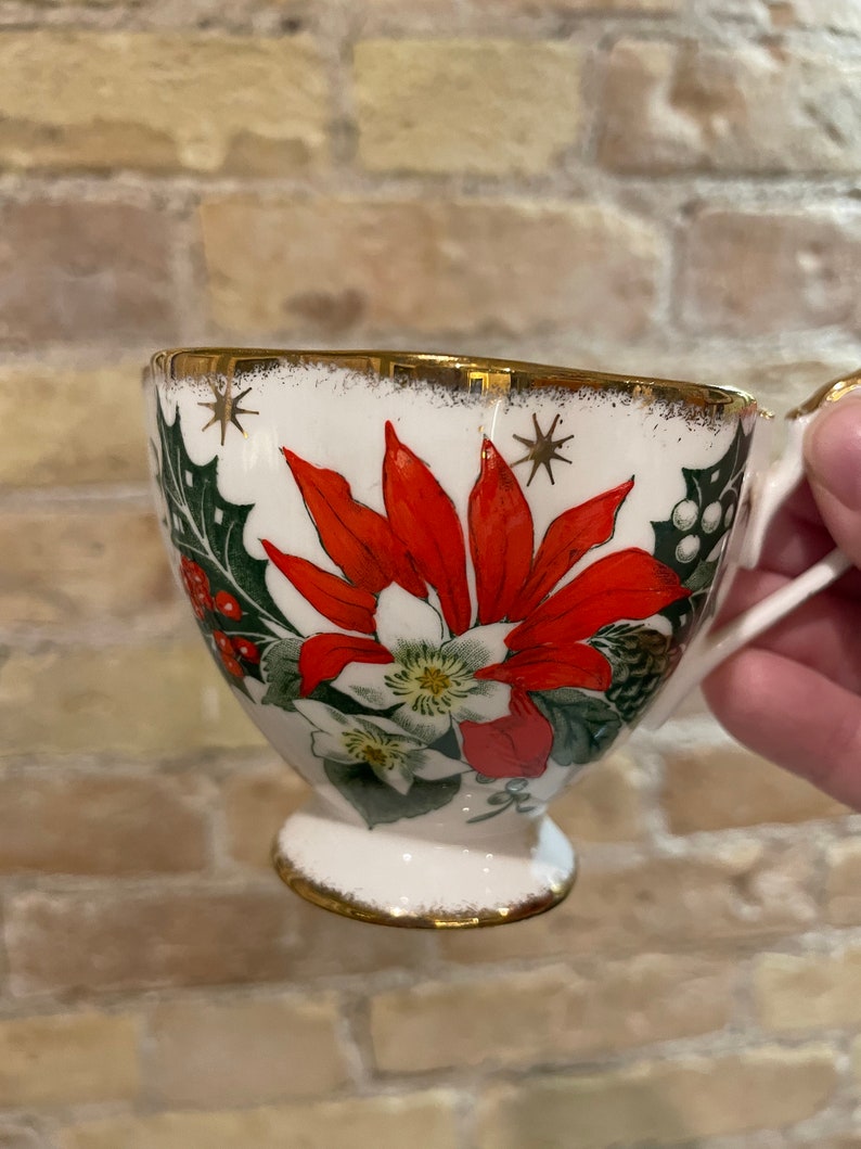 May include: White ceramic teacup with a gold rim and a Christmas-themed design featuring red poinsettias, green holly, and white flowers.