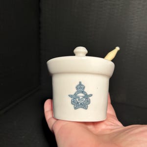 Peut inclure: Petit pot en céramique blanc cassé avec couvercle et petite cuillère. Le pot présente un emblème bleu. Le pot est tenu dans une main sur fond sombre. Le pot est probablement destiné à conserver des condiments ou de petits objets.