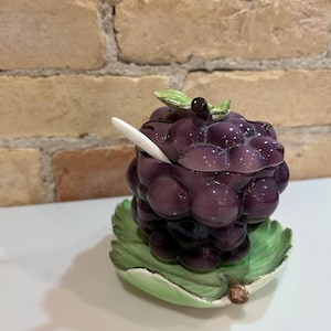 Könnte beinhalten: Eine Keramik-Zuckerdose in Form einer Weintraube mit Deckel und einem kleinen Löffel. Die Dose ist in Violett- und Grüntönen bemalt und zeigt ein realistisches Traubendesign. Die Dose steht auf einem grünen, blattartigen Sockel.