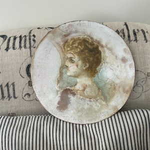 May include: A round, white plate with a painted portrait of a woman with blonde hair. The plate has a distressed, aged look and is signed "Lillian".