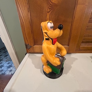 Könnte beinhalten: Eine gelbe Plastikfigur von Pluto, dem Hund aus Disney, der ein grünes Haus hält. Die Figur steht auf einem schwarzen Sockel.