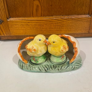 May include: A ceramic Easter salt and pepper shaker set. The set features two yellow chicks with orange beaks and accents, perched on green grass bases. They are nestled within a cracked eggshell, all resting on a green oval base.