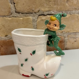 Könnte beinhalten: Ein weißer, stiefelförmiger Keramik-Blumentopf mit grünen Stechpalmen- und roten Beerenakzenten. Eine grüne Elfenfigur mit gelber Mütze und lächelndem Gesicht sitzt am Rand des Topfes. Der Topf hat eine strukturierte Oberseite.