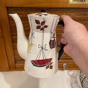 May include: White ceramic coffee pot with a black handle and lid. The pot features a design of a watermelon, bananas, and a bottle hanging from strings with bows. The design is on a white background with brown leaves.