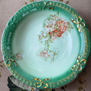 May include: A green and white porcelain bowl with gold trim and a floral design. The bowl has the text "Mich. 1904" and "Mrs. Clemens" written on it.