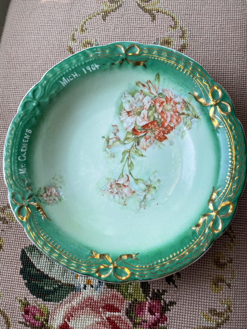 May include: A green and white porcelain bowl with gold trim and a floral design. The bowl has the text "Mich. 1904" and "Mrs. Clemens" written on it.