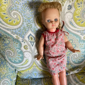 Può includere: Una bambola di plastica vintage con capelli biondi, occhi azzurri e un vestito floreale rosa e bianco. La bambola è seduta su una sedia con un rivestimento in tessuto a motivi.