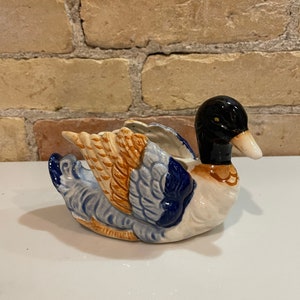 May include: A ceramic duck planter with blue, orange and white painted details. The duck has a black head and beak.