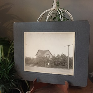 May include: A black and white photograph of a house with a porch and a garden. The house is surrounded by trees and shrubs. The photo is framed in a grey mount with a white border. The date "1904" is written in the bottom right corner of the photo.