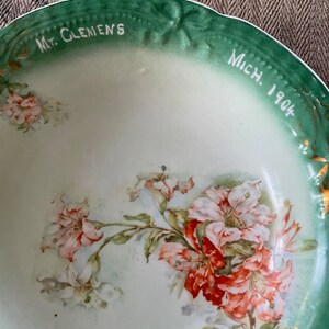 May include: A vintage green and white porcelain bowl with a floral design. The bowl has a gold rim and the words "Mt. Clemens Mich. 1904" written on the rim.