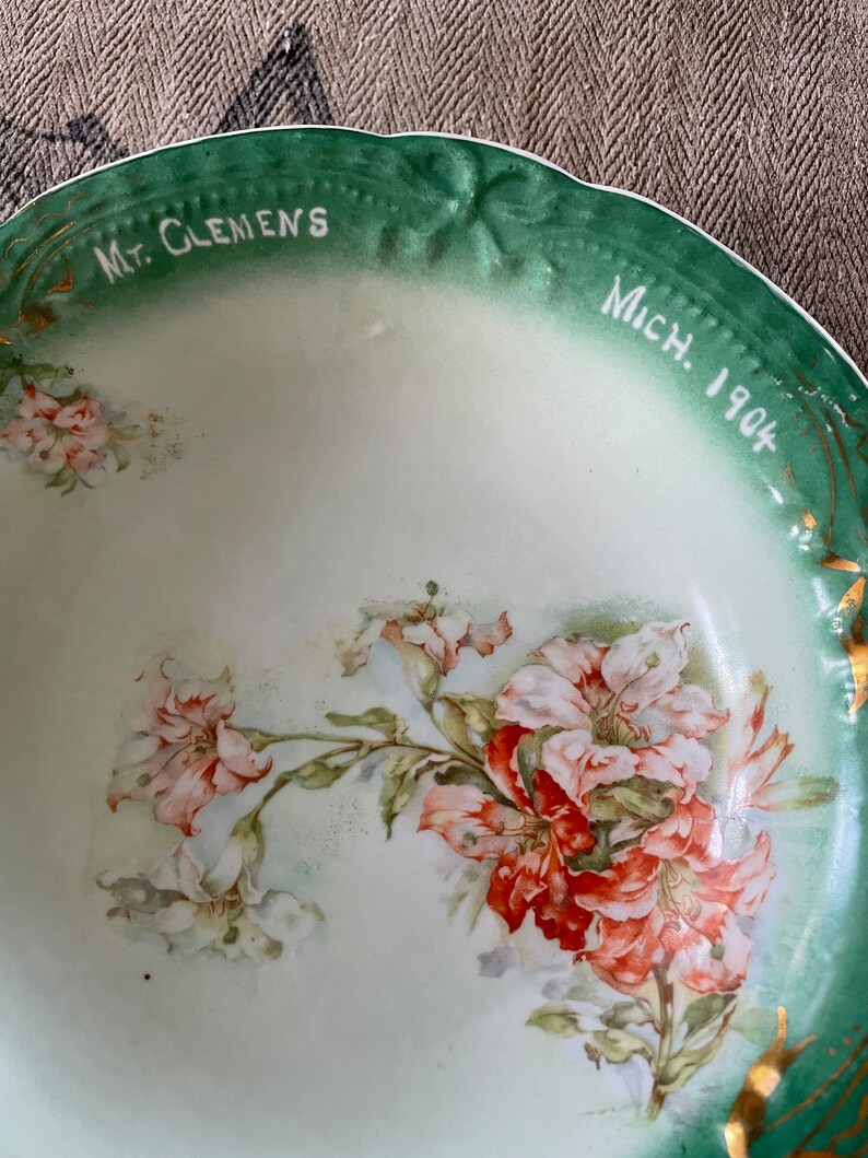 May include: A vintage green and white porcelain bowl with a floral design. The bowl has a gold rim and the words "Mt. Clemens Mich. 1904" written on the rim.