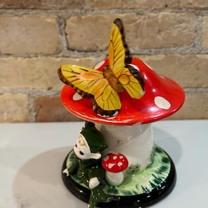 Pode incluir: Uma figurinha de cerâmica de um gnomo sentado debaixo de um cogumelo vermelho e branco com uma borboleta amarela empoleirada no topo. O cogumelo tem bolinhas brancas e o gnomo está usando um chapéu verde e uma túnica verde.