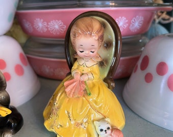 May include: A ceramic figurine of a girl in a yellow dress sitting in a chair and knitting with a cat beside her. The cat is also holding a ball of yarn.