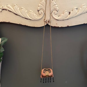 May include: A small, black and pink, beaded purse hangs from a gold chain. The purse has a gold clasp and black tassels.