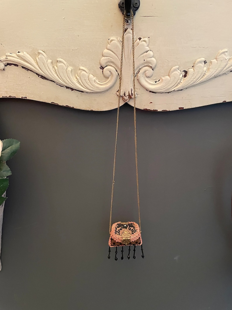 May include: A small, black and pink, beaded purse hangs from a gold chain. The purse has a gold clasp and black tassels.