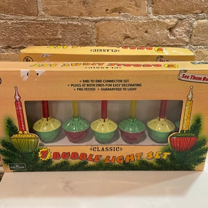 Op de afbeelding: Een doos met 7 klassieke bubbellichtjes voor kerst. De lampjes zijn rood, groen en geel. De doos is bruin met een wit label waarop "Classic 7 Bubble Light Set" staat. De doos heeft ook een UL-veiligheidslabel.