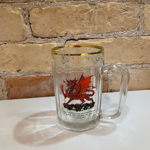 Puede incluir: Jarra de cerveza de vidrio transparente con borde dorado y asa. La jarra presenta un emblema de dragón rojo con el texto "THE INVESTITURE AT CAERNARVON OF H.R.H. THE PRINCE OF WALES 1st JULY 1969".