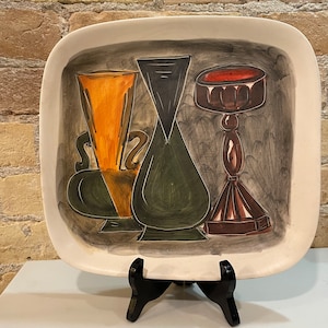 Puede incluir: Una bandeja cuadrada de servir de cerámica blanca con un diseño pintado de tres jarrones estilizados. Los jarrones están pintados en tonos de verde, marrón, naranja y negro. La bandeja está sobre un soporte negro.