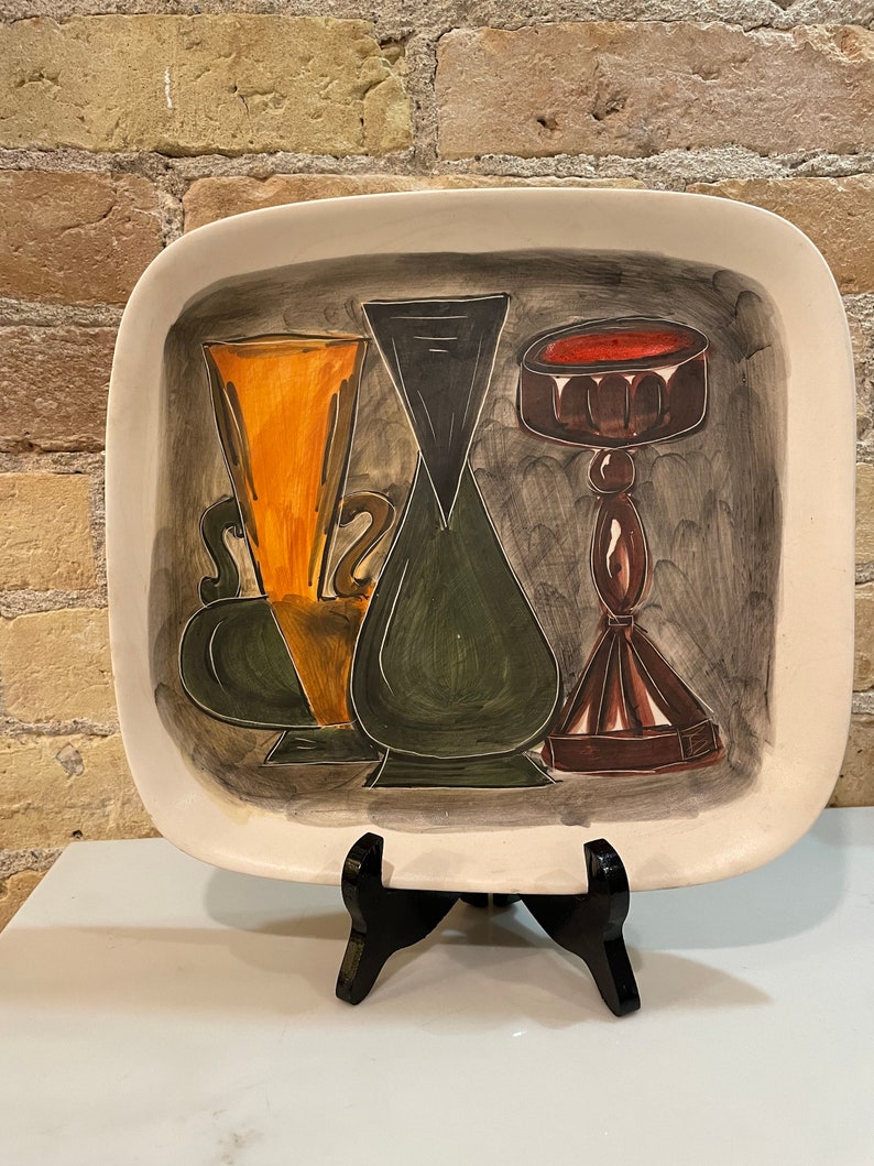 May include: A white ceramic square serving tray with a painted design of three stylized vases. The vases are painted in shades of green, brown, orange, and black. The tray is on a black stand.