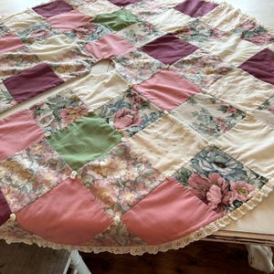 May include: A circular patchwork tree skirt featuring a variety of fabric squares in shades of pink, plum, sage green, and cream. The design includes solid colors and floral patterns, accented with small white fabric knots and a white eyelet lace trim.