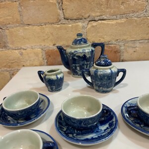 May include: A miniature blue and white porcelain tea set. The set includes a teapot, sugar bowl, creamer, and several teacups with saucers. The design features a landscape scene. The set is displayed on a white surface.