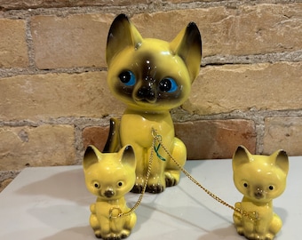 May include: A set of three ceramic Siamese cat figurines. The largest cat is sitting with a gold chain attached to its collar. The chain is connected to two smaller cats, each sitting with a gold chain attached to their collars.
