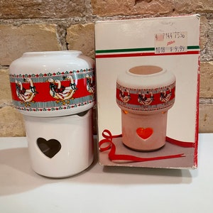 May include: White ceramic candle warmer with a red band featuring a duck design. The warmer has a heart-shaped cutout and a matching box. The box has a red, green, and white stripe design.