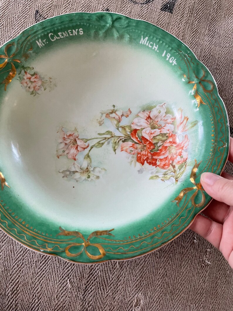 May include: A vintage green and white porcelain bowl with a floral design and gold trim. The bowl has the text "Mt. Clemens Mich. 1904" on the rim.