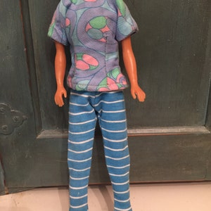 May include: A doll wearing a blue and purple patterned shirt with pink and green accents and blue and white striped pants.
