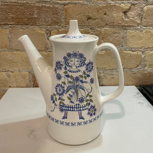 May include: White ceramic coffee pot with a blue and green floral design featuring a stylized woman. The pot has a lid and a spout.