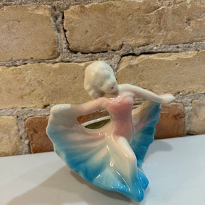 Puede incluir: Un macetero de cerámica con forma de mujer con un vestido rosa y azul. El macetero es de estilo vintage y sería una gran adición a cualquier hogar.